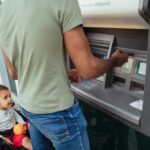 Man putting his card into an ATM machine while his son sits in a stroller beside him.