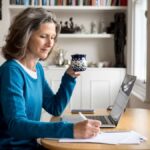 Mature Caucasian woman sat at a table with coffee and laptop while making notes on paper