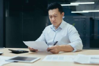 Asian man looking concerned while studying paperwork at his desk in an office