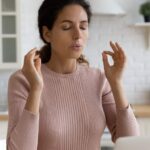 Mindful young woman breathing out with closed eyes, calming down in stressful situation, working on computer in modern kitchen.