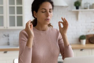 Mindful young woman breathing out with closed eyes, calming down in stressful situation, working on computer in modern kitchen.